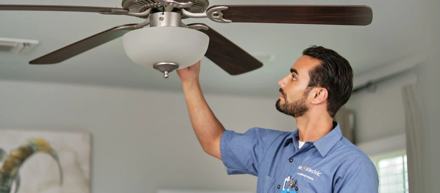 A Mr. Electric electrician reaches up to place his hand beneath the light fixture attached to a ceiling fan.