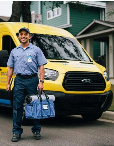 A smiling Mr. Electric electrician stands in front of a Mr. Electric van holding a bag with a rolled door mat on top of it