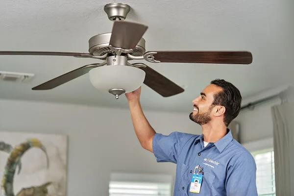 A smiling Mr. Electric electrician reaches up to the top of a light fixture attached to a ceiling fan.