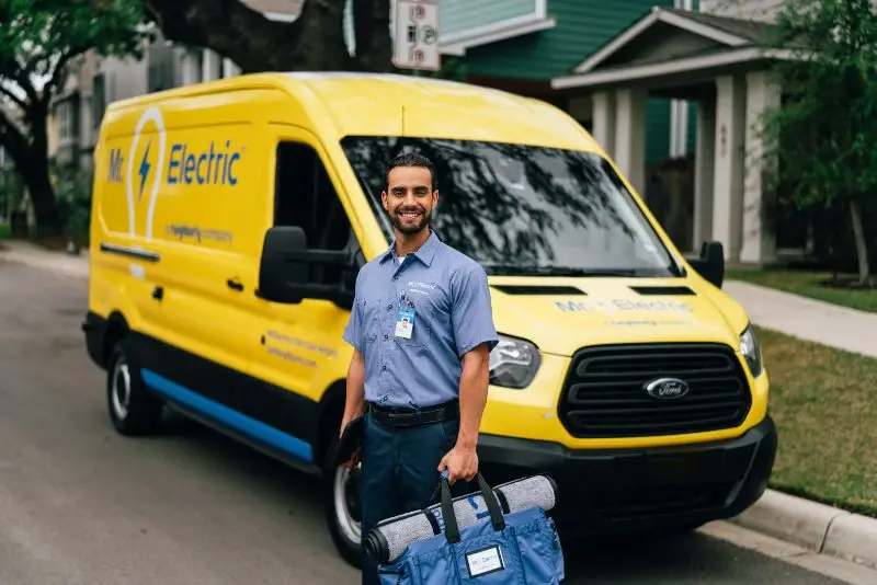 A Smiling Mr. Electric Electrician Stands in Front of a Mr. Electric Van Holding a Bag with a Rolled Door Mat on Top of It