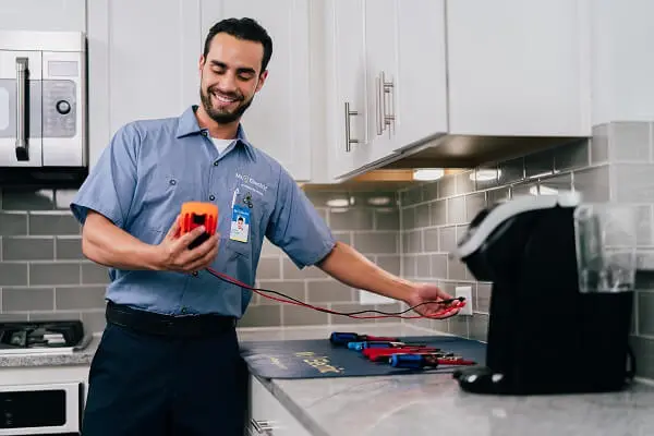 A Smiling Mr. Electric Electrician Stands Next to a Counter with Tools on it and Uses a Device to Test an Electrical Outlet