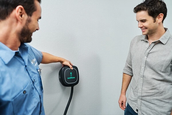 A Smiling Mr. Electric Electrician Touches an EV Charger on the Wall to Show Another Smiling Man Watching How to Use It.