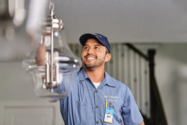 A Smiling Mr. Electric Service Professional Reaches Up to a Pendant Light Fixture Suspended from a Ceiling