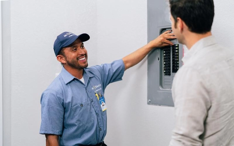 A smiling Mr. Electric electrician points at an open electrical panel while talking to a man with his back to the camera.