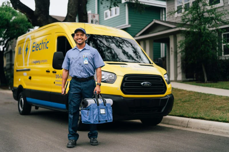 A smiling Mr. Electric electrician stands in front of a Mr. Electric van holding a bag with a rolled door mat on top of it