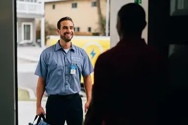 A Man in Shadow Pictured from Behind Opens the Door for a Smiling Mr. Electric Electrician Standing Outside Holding a Bag.