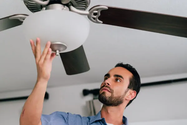 A Mr. Electric electrician reaches up to place his hand beneath the light fixture attached to a ceiling fan