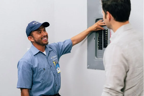 A smiling Mr. Electric electrician points at an open electrical panel while talking to a man with his back to the camera.