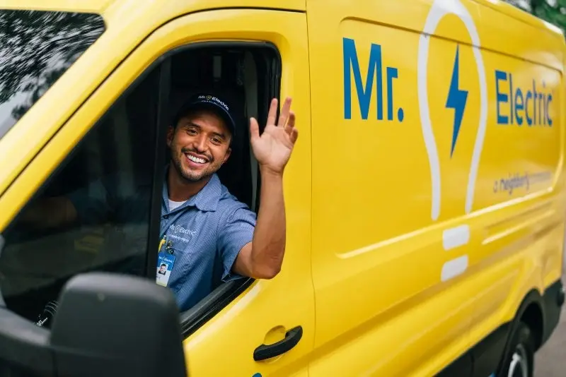 Mr. Electric professional waving and smiling from the front seat of a yellow Mr. Electric van.