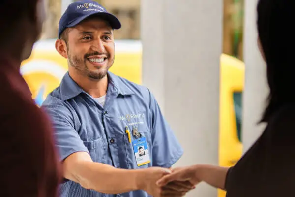 A smiling Mr. Electric electrician shakes hands with a woman with his yellow Mr. Electric van in the background behind him