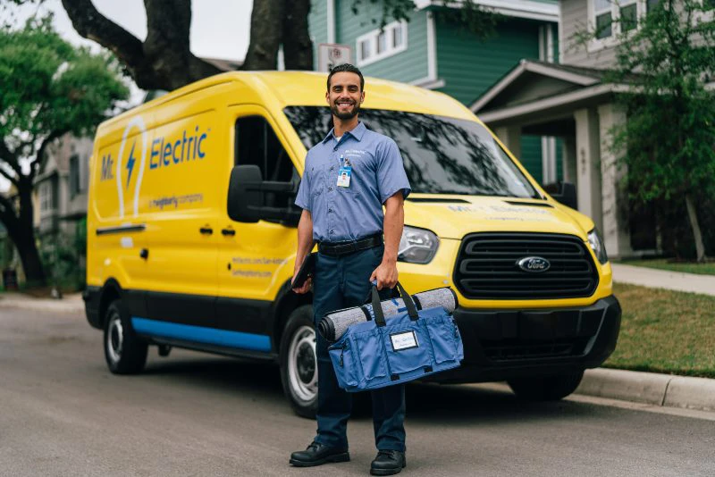 A Smiling Mr. Electric Electrician Stands in Front of a Mr. Electric Van Holding a Bag with a Rolled Door Mat on Top of It.