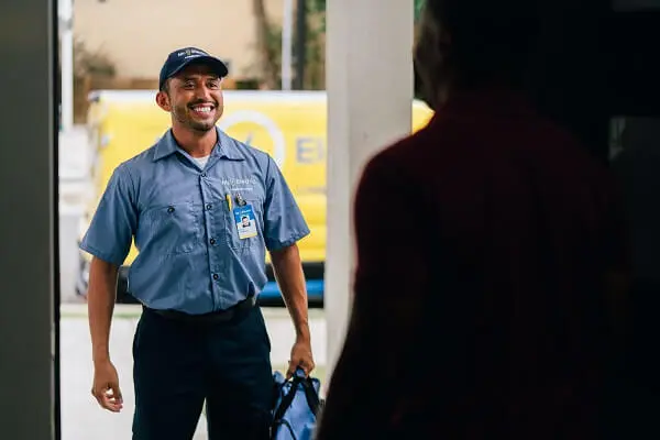 A Smiling Mr. Electric Electrician Stands in Front of an Open Door Holding a Bag While a Man Inside the Doorway Looks at Him.