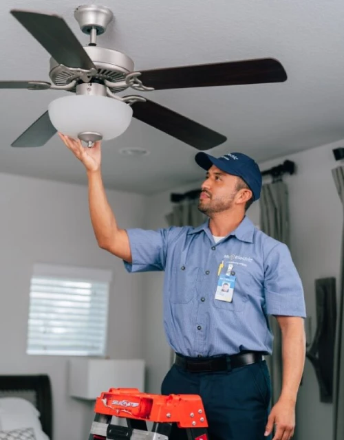 Male Mr. Electric associate in branded blue collared shirt standing on ladder and examining ceiling fan.