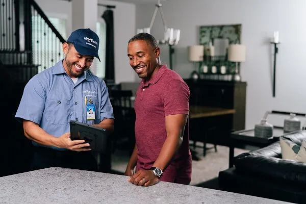 A smiling Mr. Electric electrician points at a tablet he holds as a smiling man beside him looks.