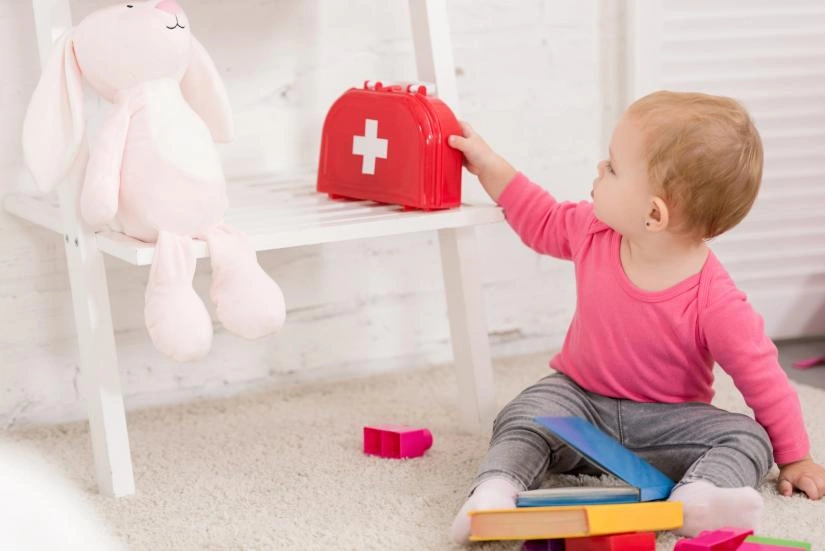 Toddler reaching for red first aid kit toy beside rabbit plushie on chair in nursery room.