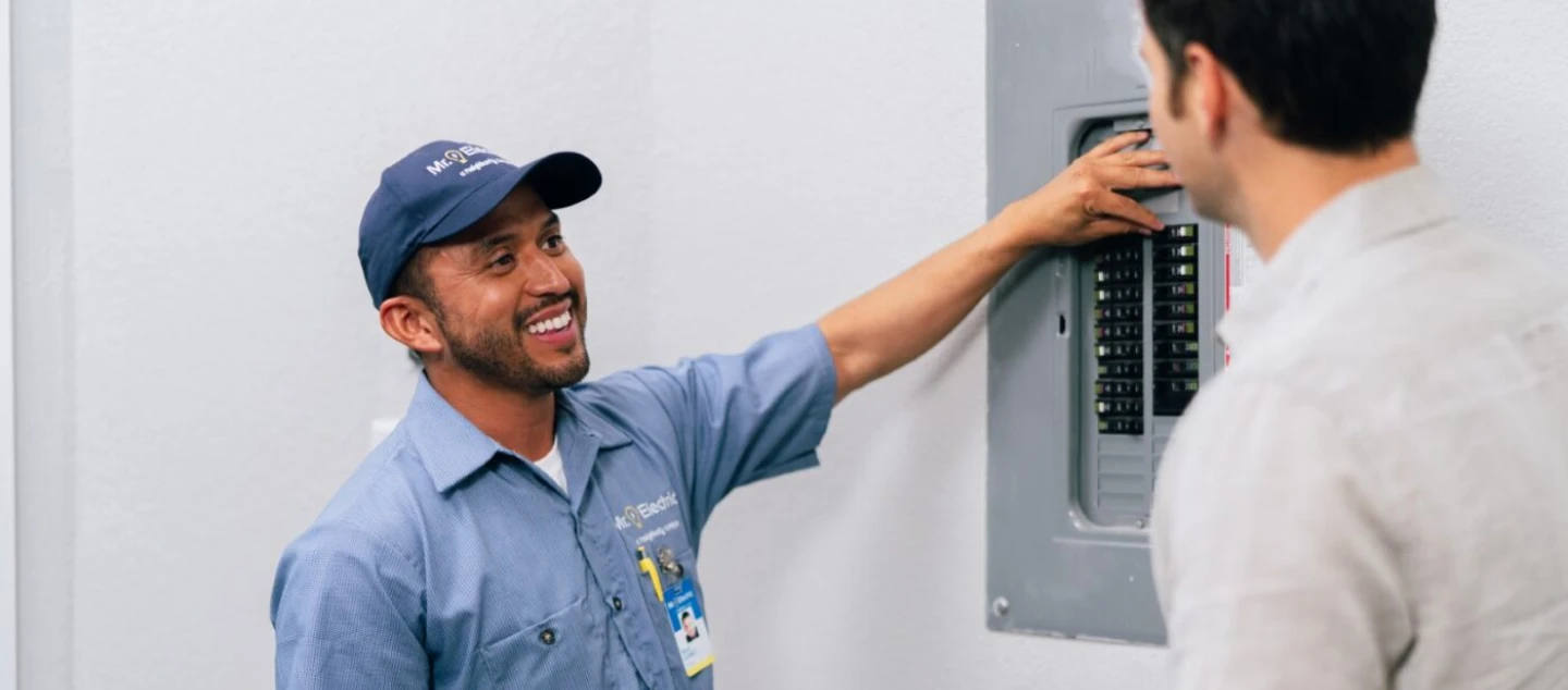 Electrician smiling and standing near circuit breaker with customer.