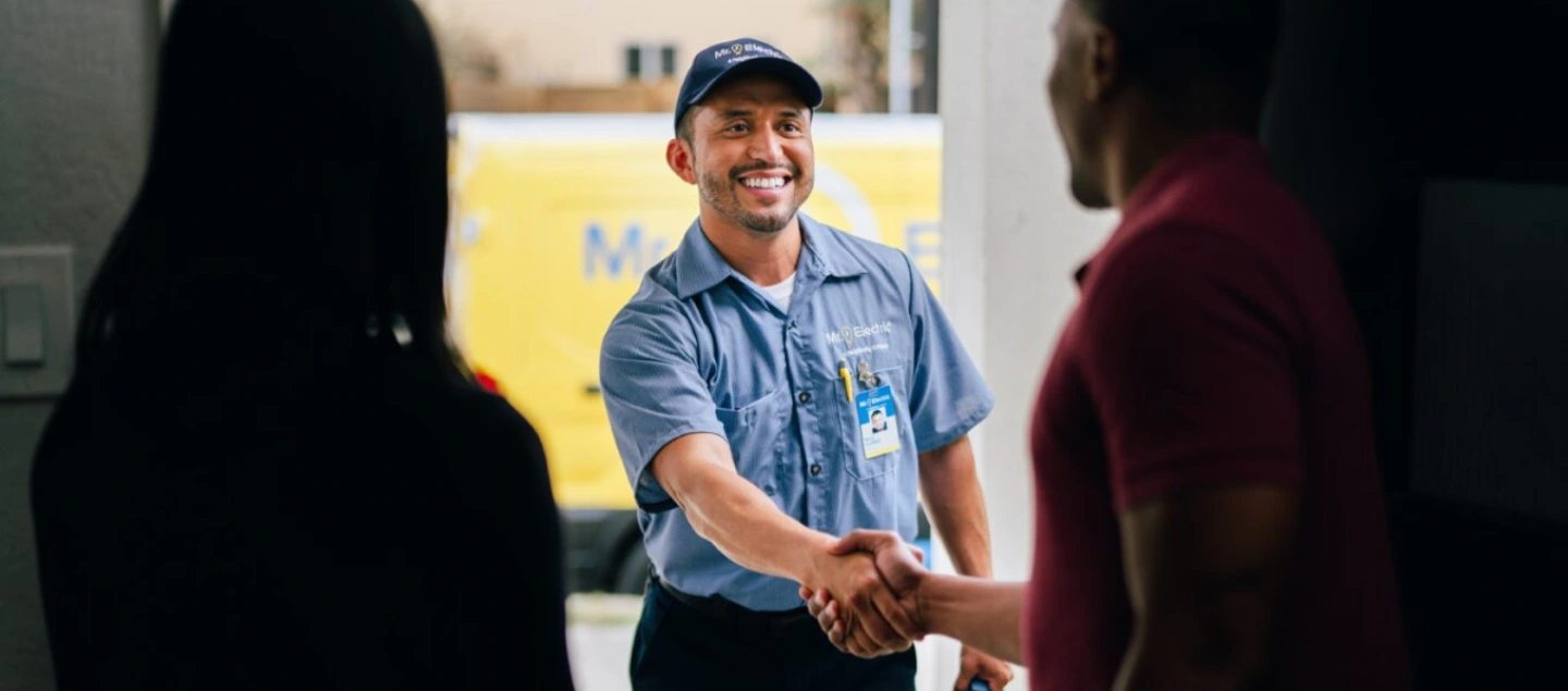 A Woman in Shadow Pictured from Behind Take a Business Card from a Smiling Mr. Electric Electrician Standing Outside Her Door