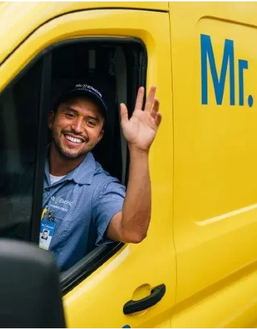 A Smiling Mr. Electric Technician in a Yellow Mr. Electrician Van Waves His Arm Out of the Open Driver-Side Window.