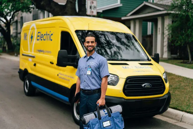 A Mr. Electric Tech Holding a Bag with a Floor Mat on it in One Hand and a Tablet in the Other in Front of a Mr. Electric Van.