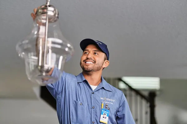 A smiling Mr. Electric electrician reaches up to a pendant light fixture hanging from a ceiling.