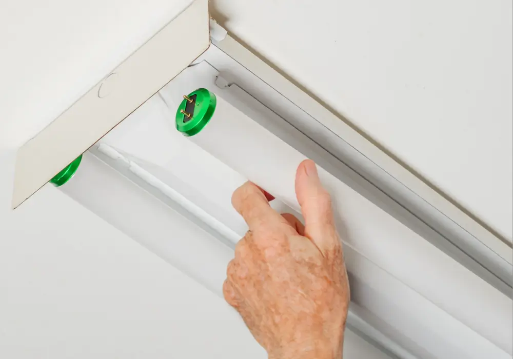 Closeup of an adult hand replacing a tubular fluorescent bulb in a white ceiling fixture with a green end cap, focusing on the details of the light and connection.