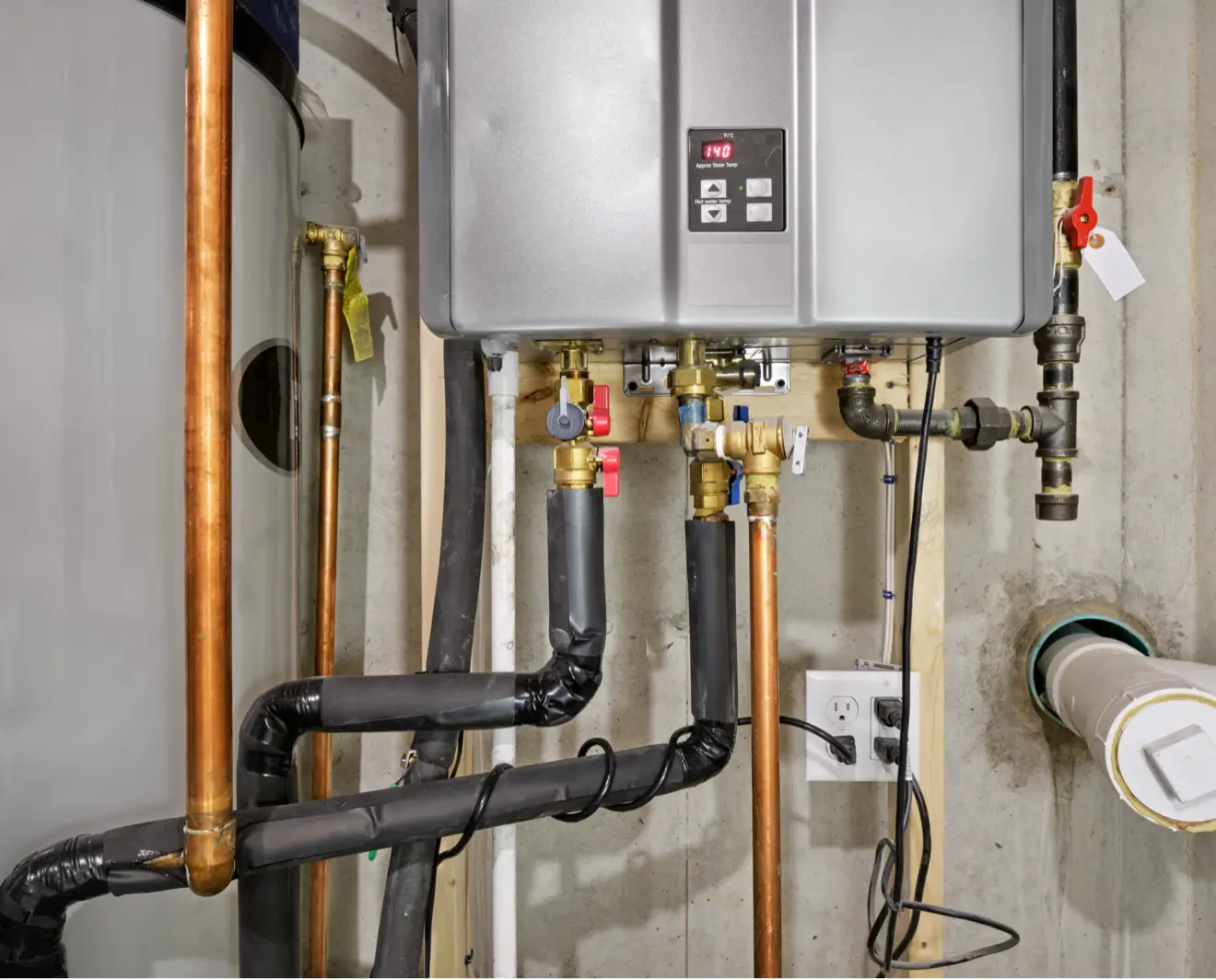 Close-up of a wall-mounted tankless water heater with metallic casing, digital display, and an array of copper and black insulated pipes connected to the system on a concrete basement wall.