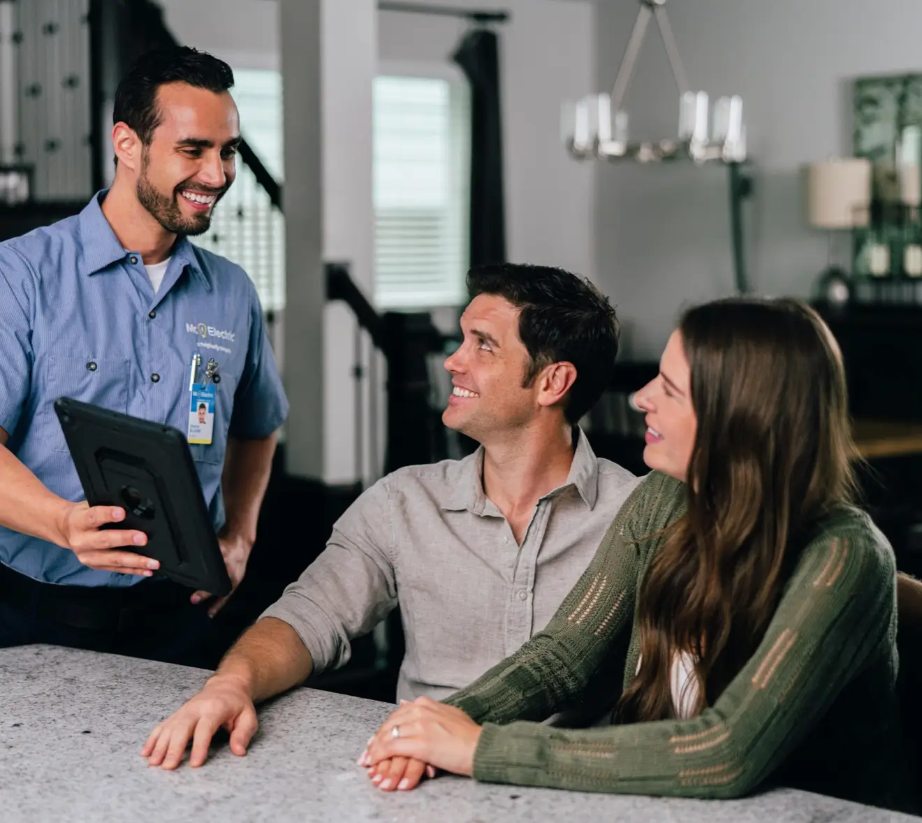 Smiling electrician in a blue shirt holding a tablet and discussing electrical services with a man and woman seated together at a marble kitchen island in a modern, open-concept home.