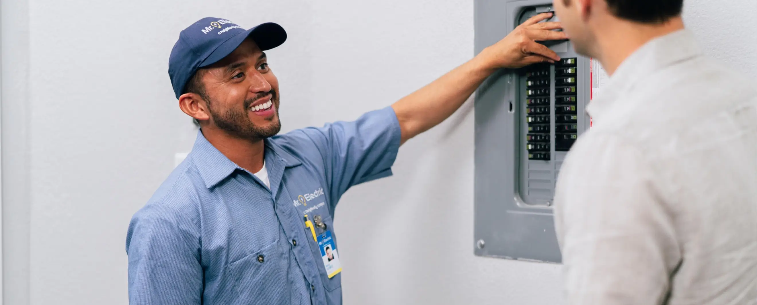Electrician in a blue uniform smiling and explaining the contents of a gray circuit breaker panel to a homeowner whose back is to the camera, inside a white room.
