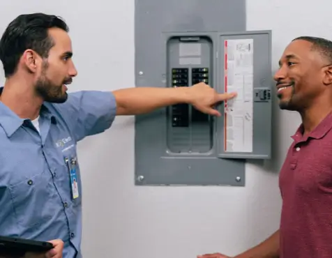 Electrician in a blue uniform pointing at the breakers and chart inside an open electrical panel while talking with a smiling customer in a maroon shirt in a white-walled room.