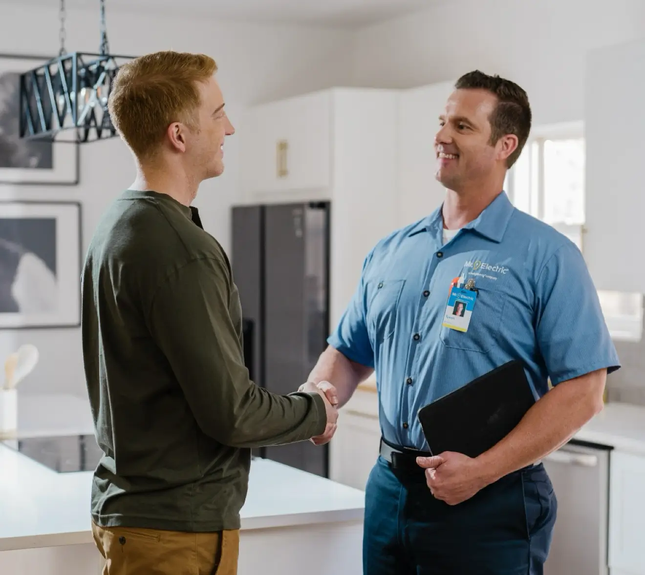 Electrician in blue Mr. Electric uniform smiling and shaking hands with a customer in a bright, modern kitchen.