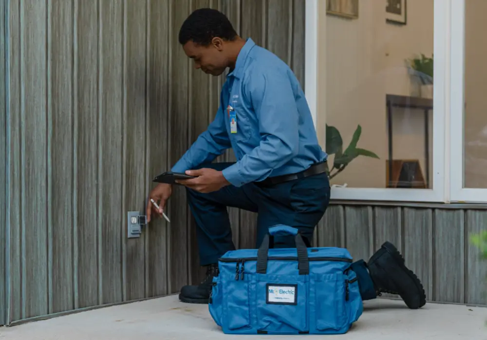 Electrician in blue uniform kneeling outdoors, inspecting or testing a wall-mounted electrical outlet with tablet and pen, blue tool bag on the ground nearby.
