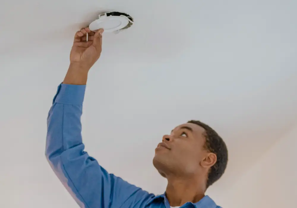 Electrician wearing a blue shirt installing or checking a round LED light fixture in a white ceiling, looking up while reaching overhead.