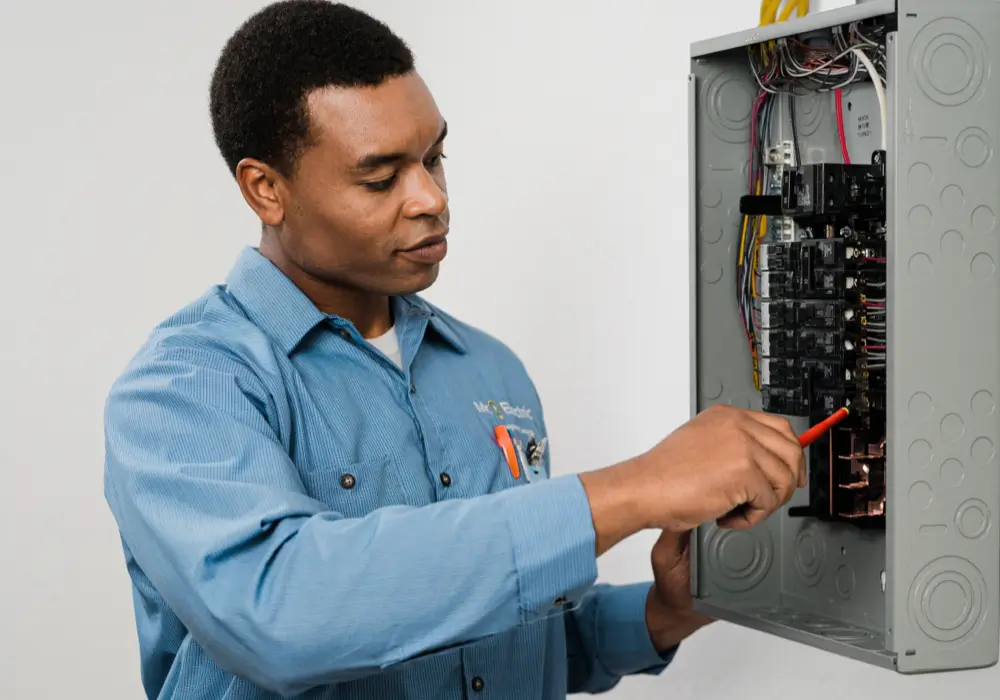Electrician in a blue uniform working with a screwdriver on an open electrical subpanel box, showing wires and breaker switches in a clean indoor setting.