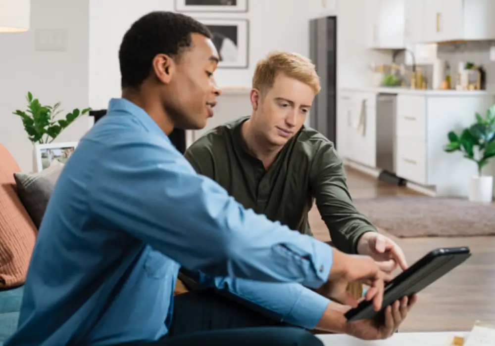 Electrician in blue shirt sitting with customer on a couch, both looking at and interacting with a tablet, in a modern, well-lit home interior setting.
