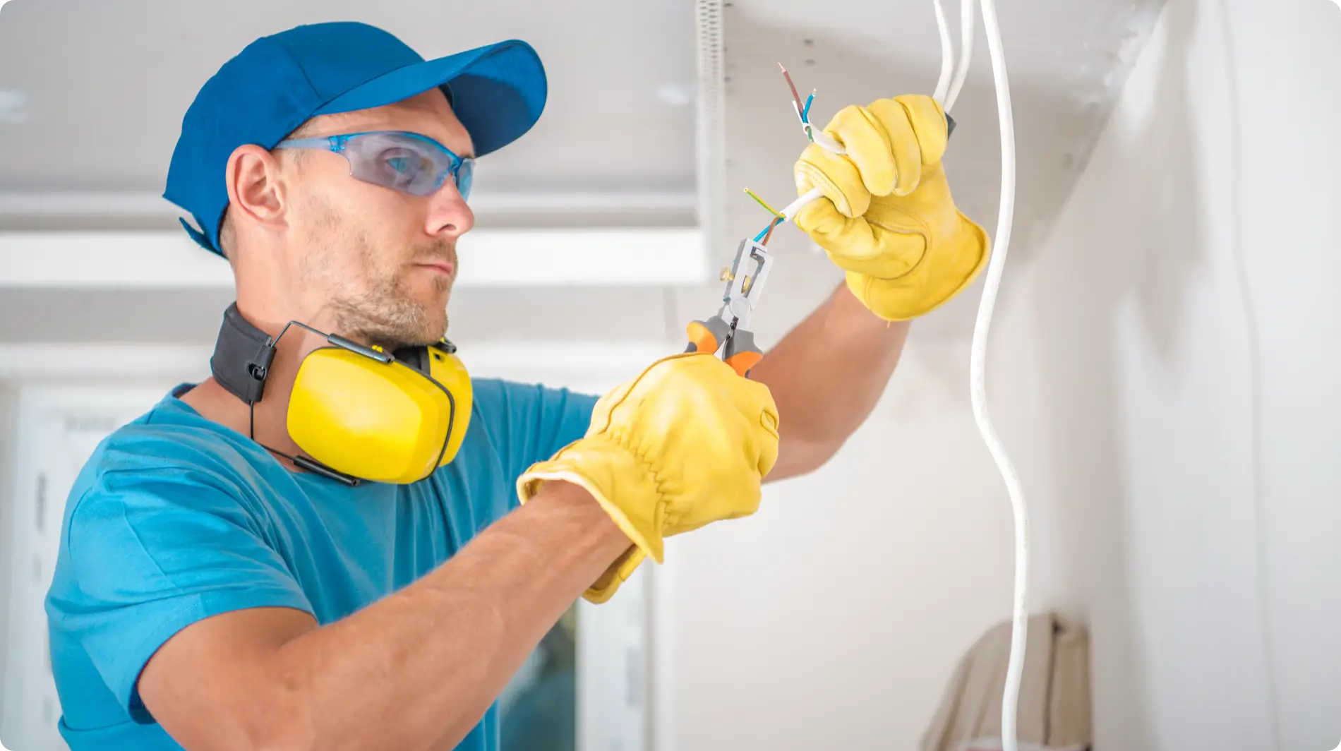 A focused electrician wearing a blue baseball cap, blue-tinted safety glasses, and yellow earmuffs is stripping the insulation from white electrical wires using orange and black wire strippers. He is wearing yellow leather gloves and a light blue T-shirt. The background is a slightly out-of-focus white interior wall.
