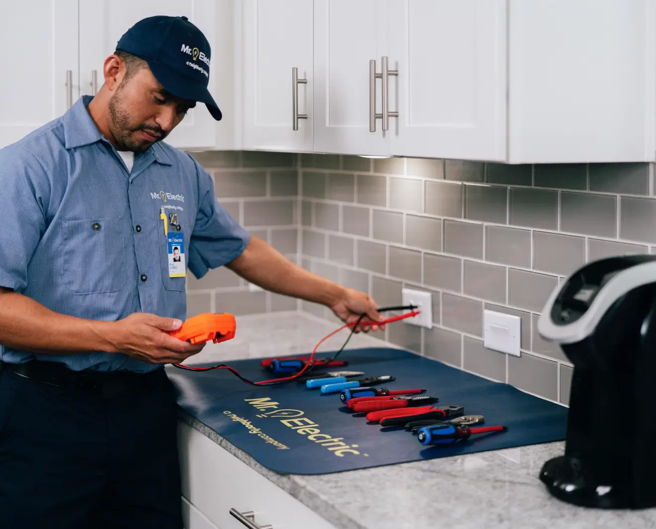 Electrician with Mr. Electric hat and blue shirt using a voltage tester to check a wall outlet on a gray tiled backsplash, with an array of electrical tools placed on the counter.