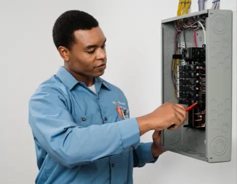 Electrician in blue Mr. Electric shirt using a screwdriver on a gray circuit breaker panel mounted on a white wall with organized yellow and blue wire bundles above the box.