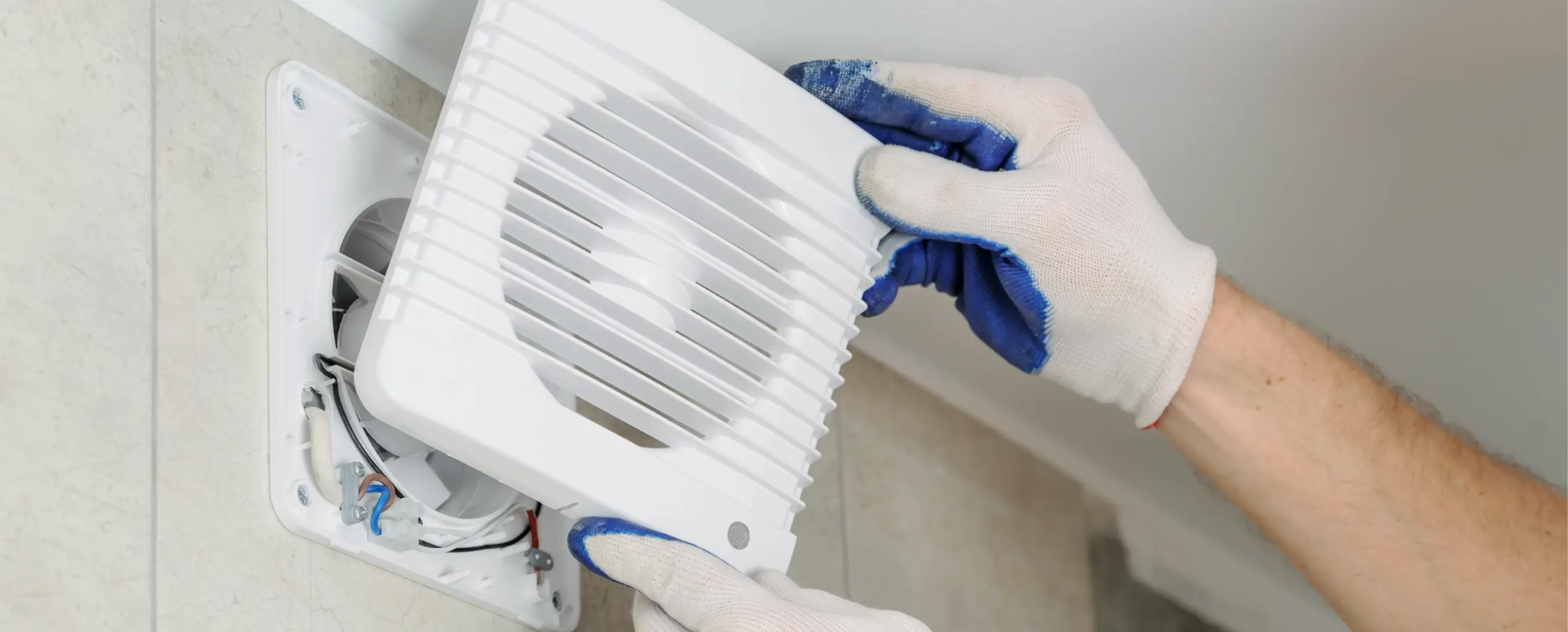 Closeup of gloved hands installing the front cover on a white ceiling exhaust fan, with visible wires and vent structure, on a tiled wall.