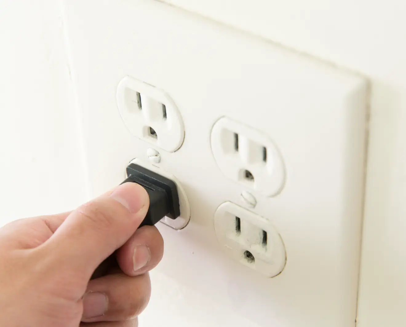 Close-up of a hand plugging a black electrical cord into a white quad wall outlet on a clean indoor wall.