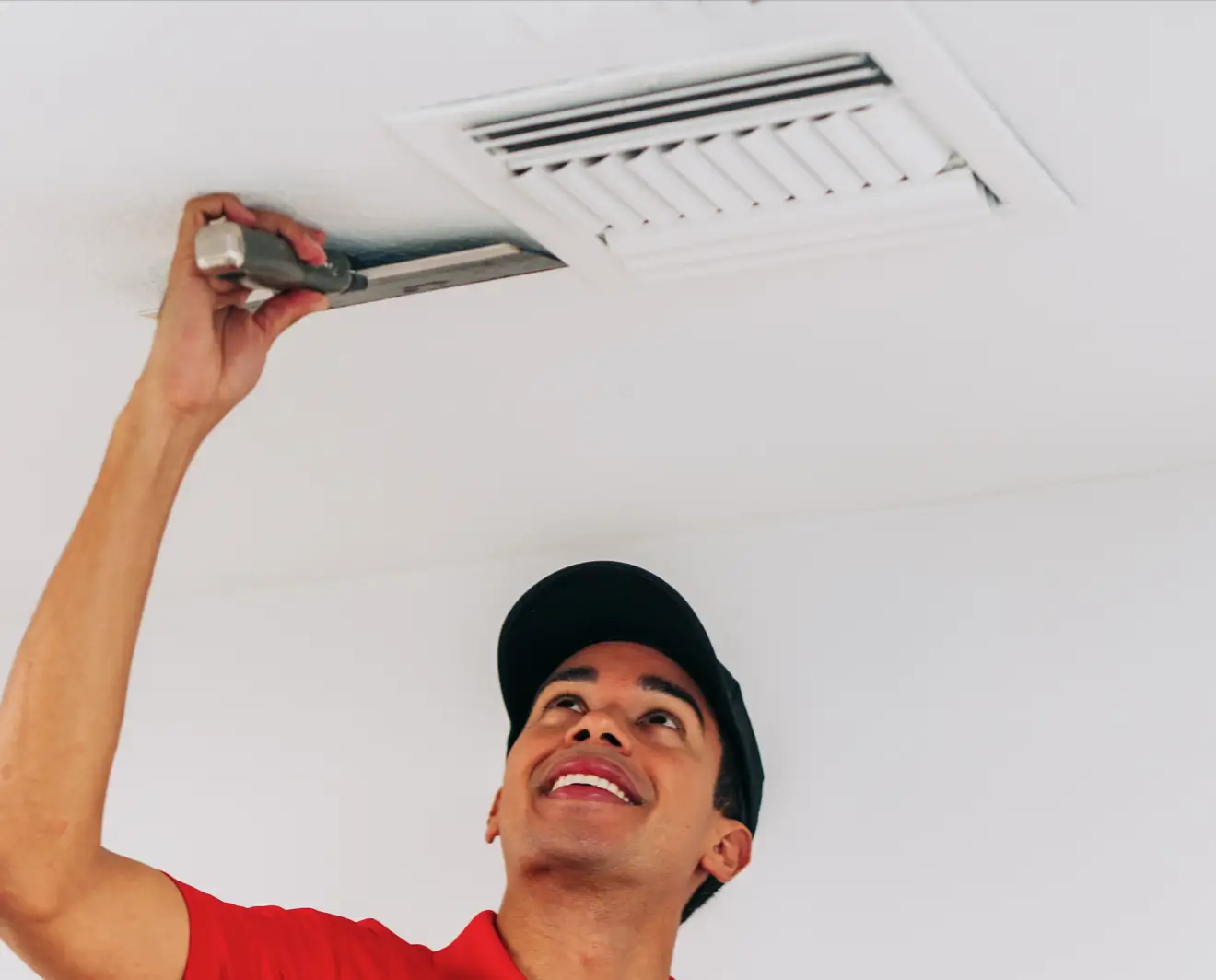 Young handyman in a red uniform shirt and black cap, using a screwdriver to clean or install a rectangular ceiling vent in a brightly lit room.