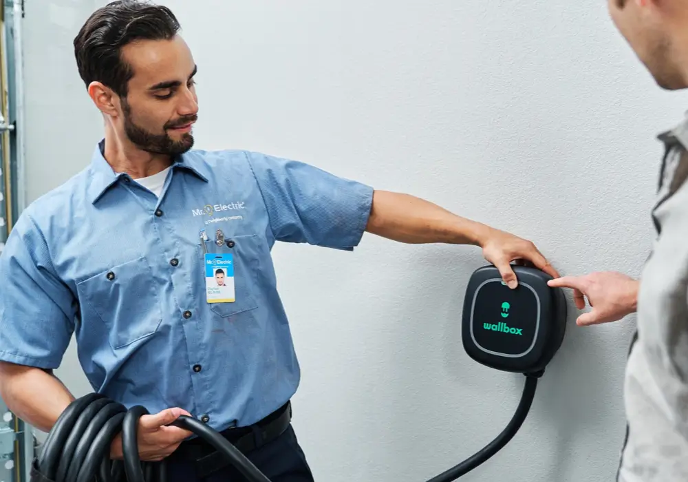 Technician in a blue work shirt and name badge demonstrating the installation of a black Wallbox electric vehicle charger mounted on a white wall, holding a charging cable and talking to a homeowner.