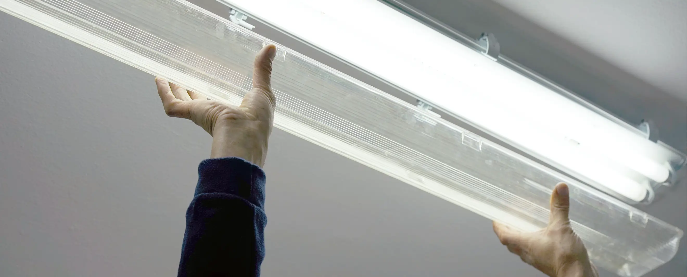 Close-up of two hands mounting or removing a transparent plastic cover from a fluorescent ceiling light fixture, with focus on the installation process.
