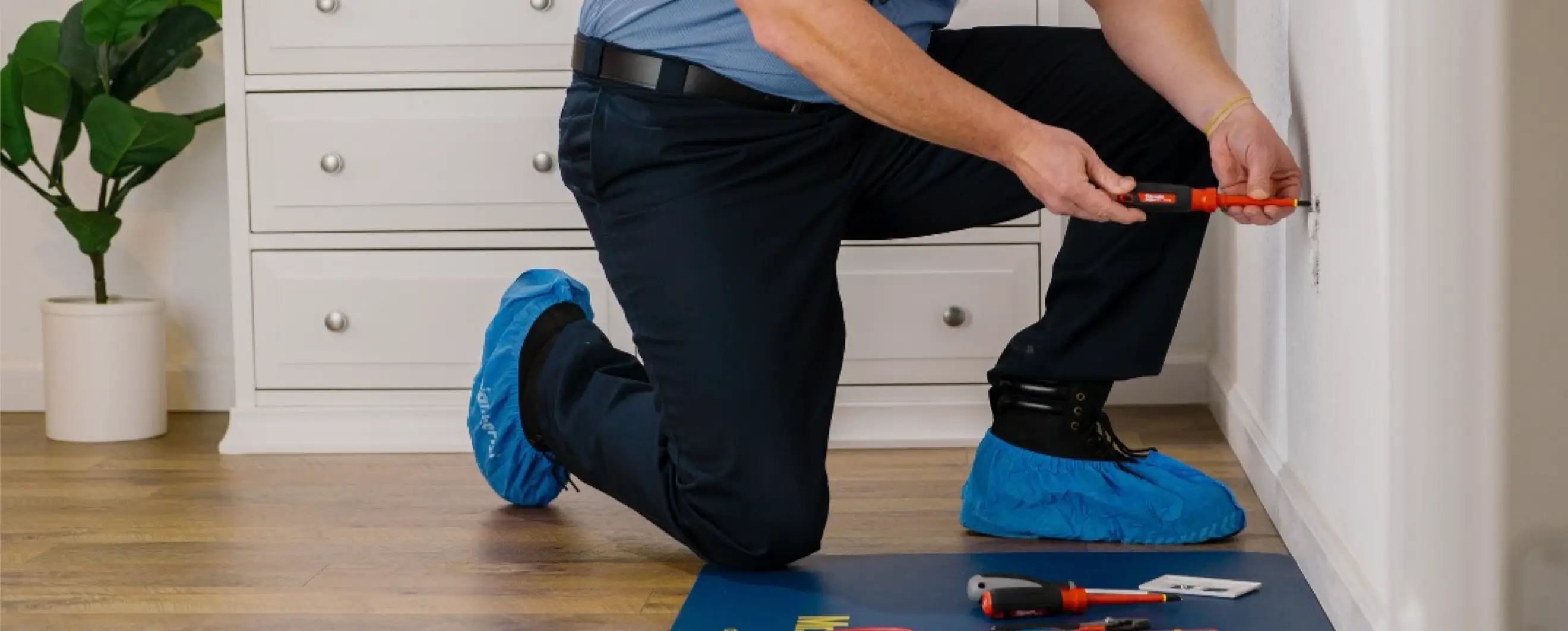 Electrician kneeling on the floor with tool bag and blue shoe covers, using a screwdriver to work on a wall outlet in a residential setting.