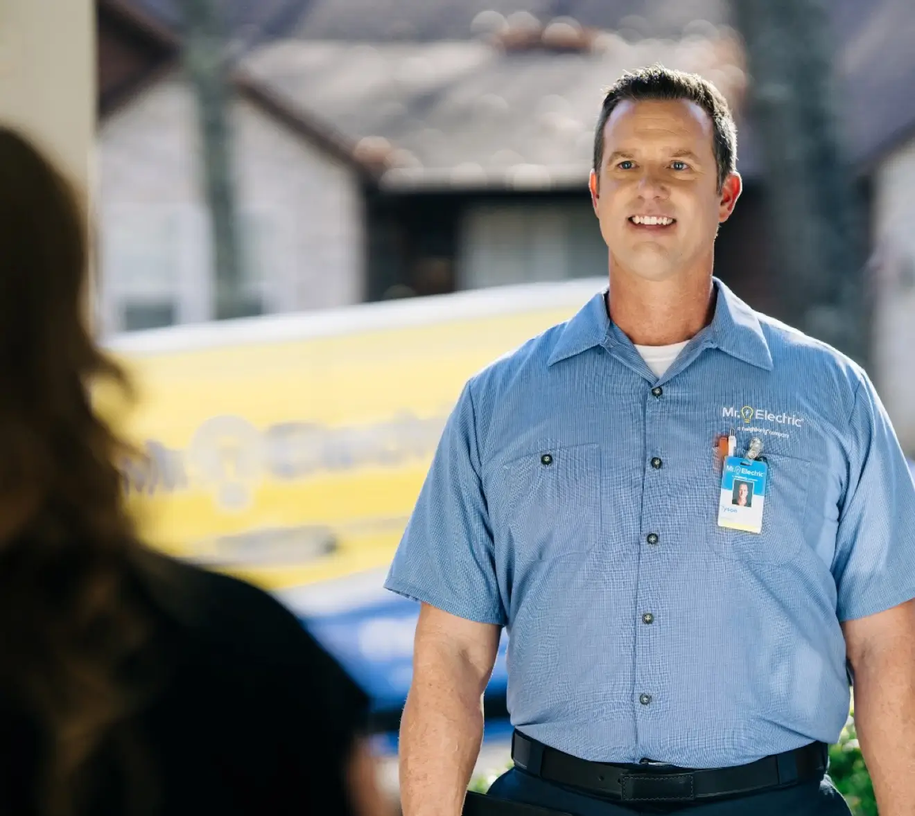 Smiling service professional in blue 'Mr. Electric' shirt with name tag, standing at a residential front door, greeting a customer with a branded van blurred behind.