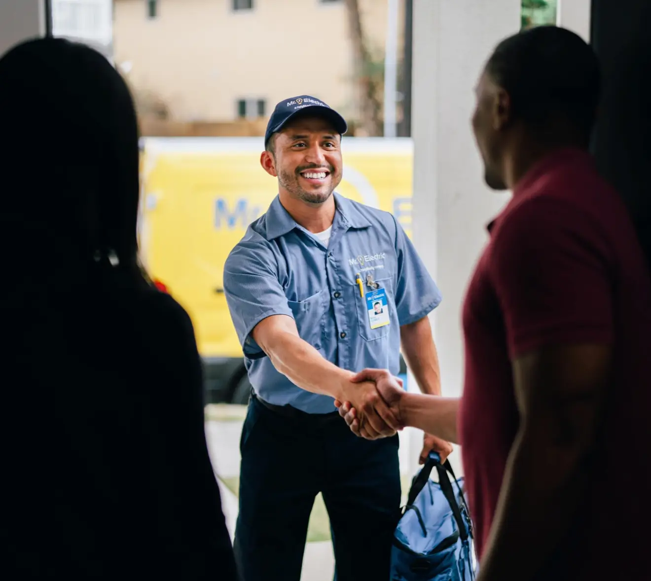 Smiling electrician in blue uniform and cap, holding a toolbag and shaking hands with homeowner at the front door, with a work van visible in the background.