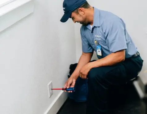 Service professional in blue uniform and cap kneeling beside a wall, using a screwdriver to test a white electrical outlet in the corner of a bright room with a window and dark flooring.