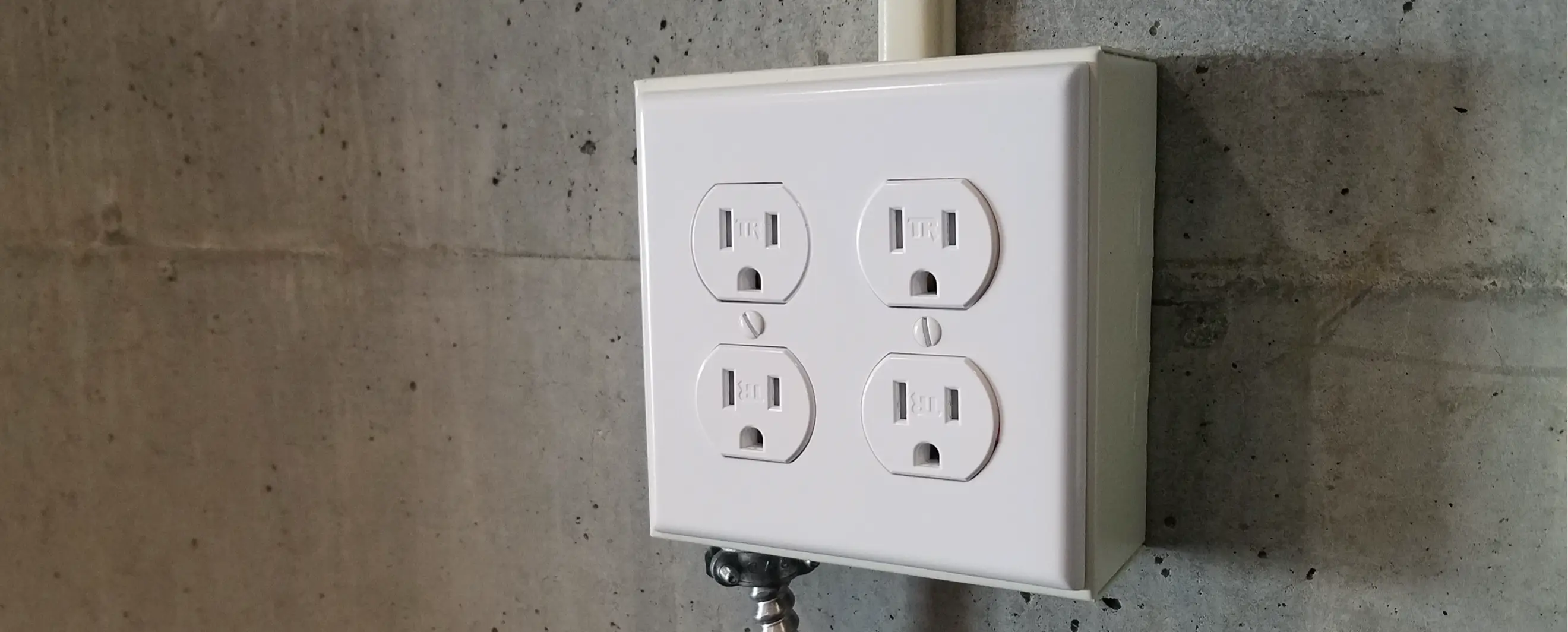 White metallic quad electrical outlet box installed on a gray concrete wall, with conduit extending upwards, shown in a utility or garage space.