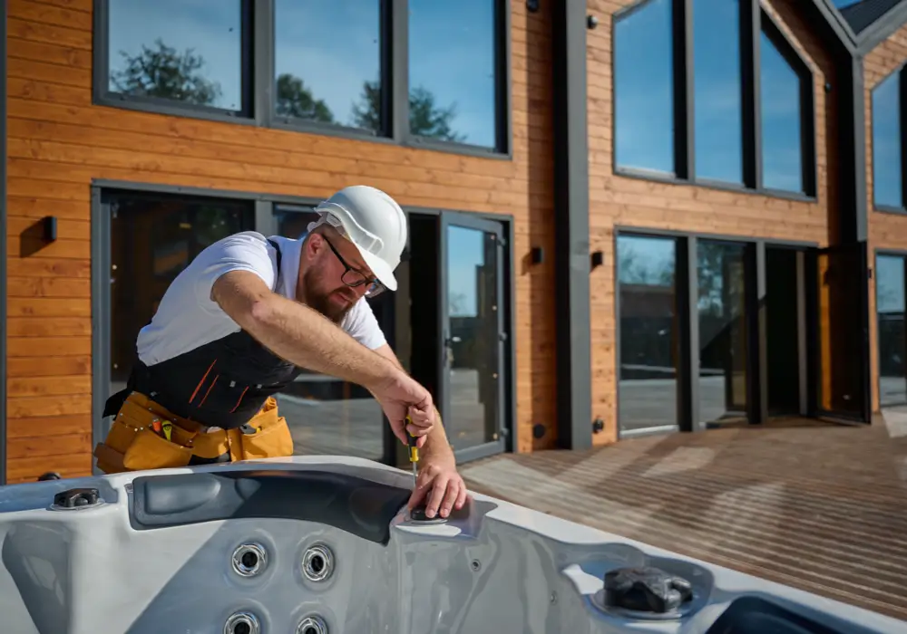 Bearded male installer in safety glasses, hard hat, and tool belt using a screwdriver on an outdoor hot tub, with wooden modern house and deck background.