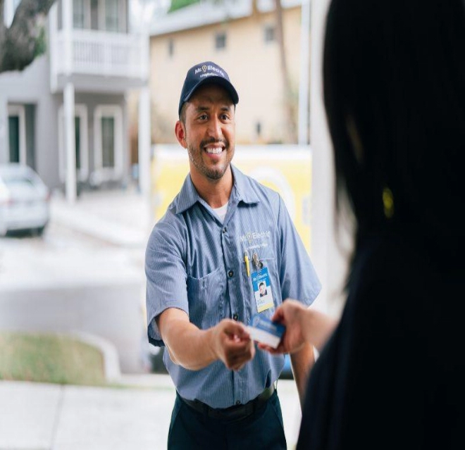 A Woman in Shadow Pictured from Behind Take a Business Card from a Smiling Mr. Electric Technician Standing Outside Her Door.