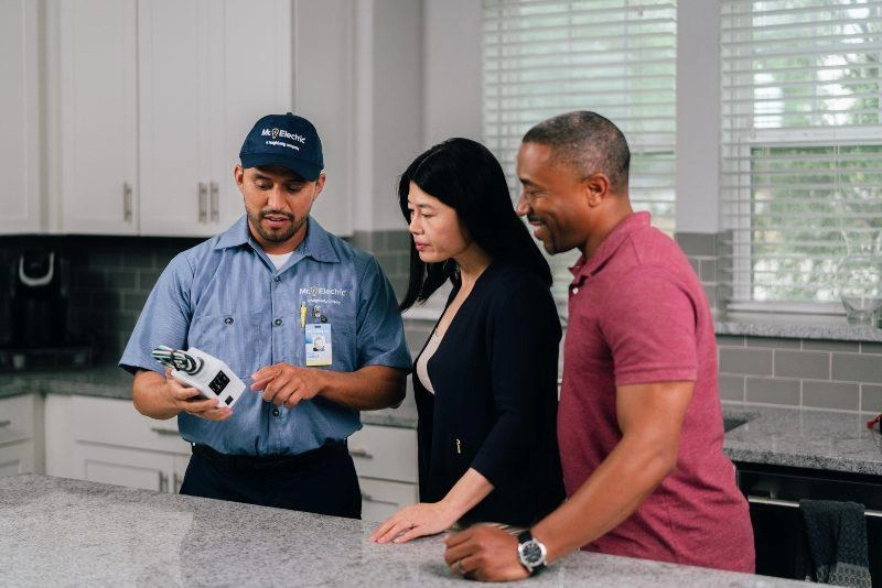 A Mr. Electric Electrician Holds and Points at a House Surge Protector and Explains it to a Woman and Man Next to Him.